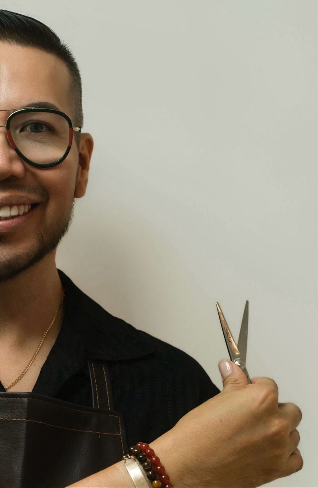 Moses smiling, wearing glasses and his studio apron, holding a pair of scissors against a light studio wall