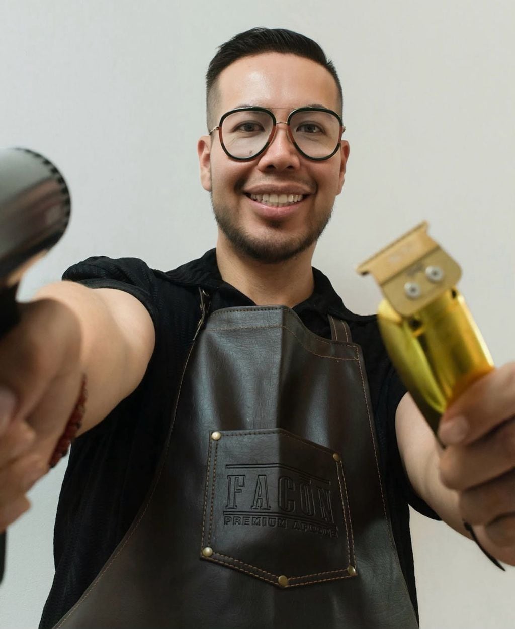 Moses smiling at the camera, holding a hair dryer and a gold trimmer, wearing his leather studio apron